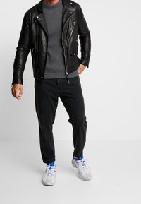 Schwarze Leder-Bikerjacke, grauer gerippter Pullover, schwarze Jeans und weiße Sneaker mit blauen Akzenten. Die Jacke verfügt über Reißverschlusstaschen.