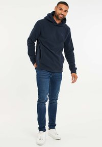 Donkerblauwe fleece hoodie met een kangoeroezak en een verstelbare capuchon, gedragen met slim-fit blauwe jeans en witte sneakers.