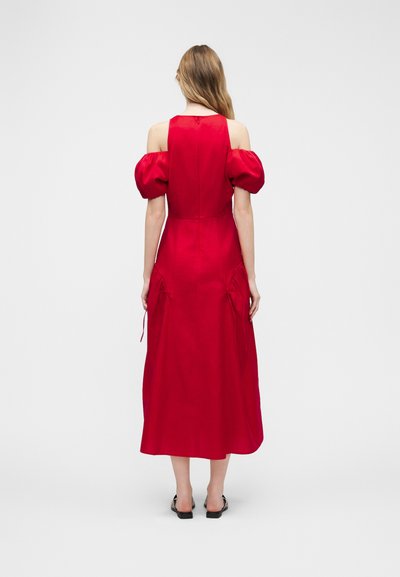 Γυναίκα που φοράει ένα κόκκινο μεσαίο φόρεμα με φουσκωτά μανίκια χωρίς ώμους και λεπτομέρειες με ρυθμιζόμενο κορδόνι, στέκεται πλάτη σε λευκό απλό φόντο.