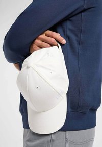 Biała czapka bejsbolowa trzymana w ręku, wykonana z materiału z przeszytymi akcentami. Posiada okrągłą koronę i zakrzywiony daszek.