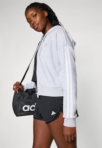 Lys grå zip-up hettegenser med hvite striper på ermene, båret over en svart topp og kombinert med svarte shorts; holder en svart bag med logo.