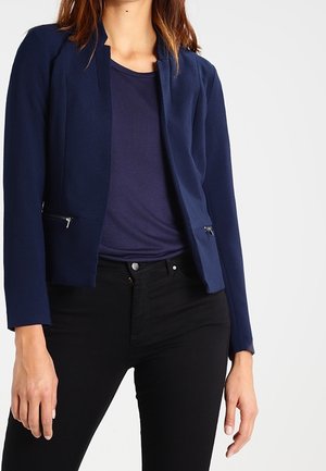 Vrouw draagt een marineblauwe blazer met ritssluitingen over een marineblauw T-shirt en zwarte strakke broek, staand tegen een effen achtergrond.