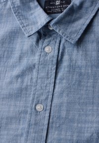 Ljusblå, kortärmad skjorta med knappstängning gjord av ett mjukt, strukturerat tyg. Har en spetsig krage och klara knappar. Etikett synlig inuti.