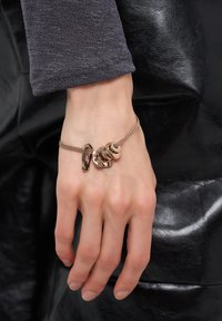 Hand med ett armband i roséguld som har abstrakta metallcharmar mot en svart läderbakgrund och mörkt långärmat tyg.