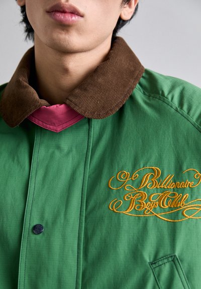 Grøn jakke med en brun kordfløjskrave, lyserød inderkrave og gul broderet "Billionaire Boys Club" logo. Har en trykknap.