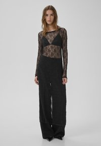 Top de encaje negro de manga larga con un diseño de escote en V profundo, superpuesto a una falda negra ajustada. Textura transparente con mangas largas, acentuando una silueta elegante.