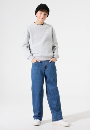 Sweat-shirt gris avec des poignets côtelés, associé à un jean bleu décontracté et des baskets blanches. Un bonnet noir complète la tenue.