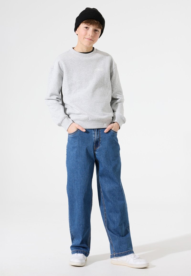 Sudadera gris con puños acanalados, combinada con vaqueros azules holgados y zapatillas blancas. Un gorro negro completa el conjunto.