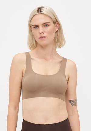 Femme blonde aux yeux bleus portant un soutien-gorge de sport beige sans couture et un bas marron foncé, se tenant devant un fond blanc uni.