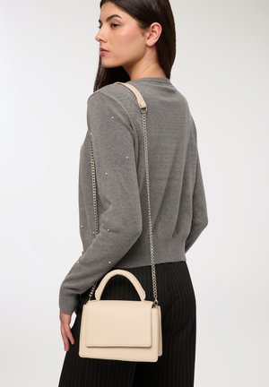 Borsa beige con un design strutturato, manico superiore e tracolla a catena. Realizzata in materiale liscio, mostrata con un maglione grigio e pantaloni a righe.