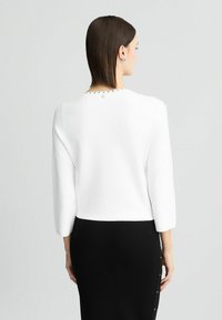 Top a maglia bianco, corto, con maniche a tre quarti. Sul retro presenta discreti borchie argentate lungo il collo. Abbinato a una gonna nera.