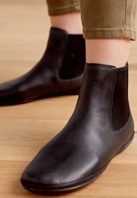 Botas de cuero negras hasta el tobillo con paneles laterales elásticos, punta redondeada y un acabado liso. Colocadas sobre un suelo de madera clara, con pantalones verdes encima.