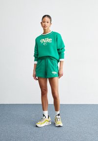 Žena stojí na modrém koberci, má na sobě zelenou mikinu Nike a šortky, bílé ponožky a žluto-bílé tenisky, proti bílé zdi.