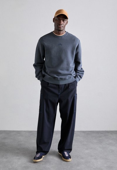 Sweatshirt azul escuro com um pequeno logotipo, combinado com calças cargo azul-marinho com costuras azul-claro e ténis bege. Boné castanho na cabeça.