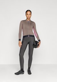 Abbigliamento da ciclismo con una maglia aderente marrone con dettagli rosa e maniche lunghe, abbinata a leggings neri. Il modello tiene un casco nero.