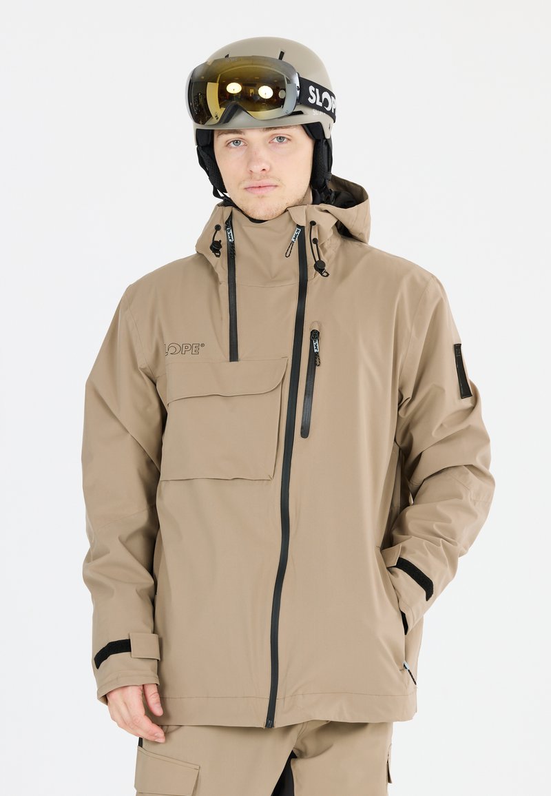 Beige vattentät skidjacka med dragkedja och flikfickor, justerbar huva och elastiska ärmmuddar. Bärs med matchande beige skidbyxor och hjälm.