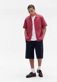 Rosa kortärmad skjorta med knappar och bröstficka, kombinerad med mörka randiga shorts och bruna snörskor. Vita strumpor syns.