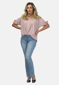 Jasnoróżowa bluzka z krótkim rękawem, z przodu skręcona i z akcentami w postaci kokard na rękawach, w połączeniu z jasnoniebieskimi dopasowanymi dżinsami i czarnymi szpilkami.