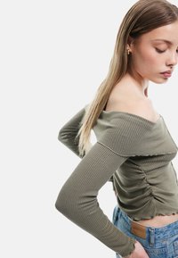 Khakifarbenes off-shoulder Ripp-Oberteil mit langen Ärmeln, das über geraffte Details an der Taille und gewellte Säume verfügt. Sichtbare Jeans aus Denim.