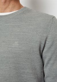 Grauer Strickpullover mit einem strukturierten Muster, runder Halsausschnitt und dezente Logo-Details auf der Brust. Material aus Baumwollmischung.