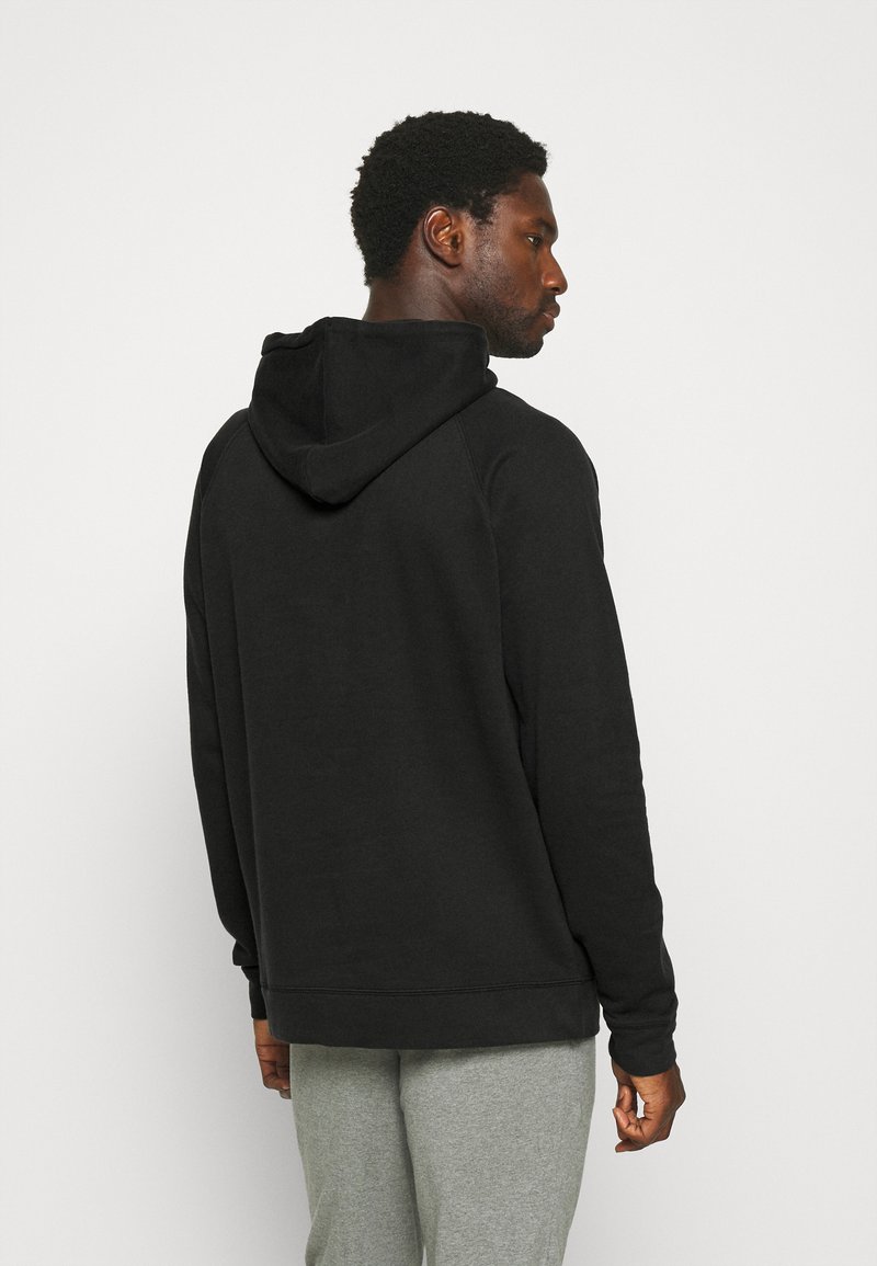 Schwarzer Kapuzenpullover mit weicher Textur, lockerer Passform und gerippten Bündchen, über grauen Hosen getragen. Die Rückansicht hebt die Form der Kapuze hervor.