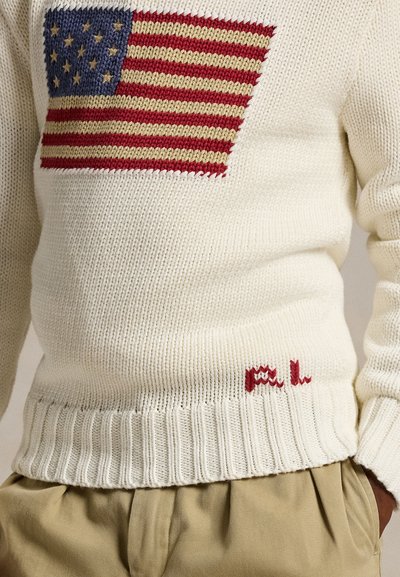 Valkoinen neulottu villapaita, jossa on neulottu amerikkalainen lippu rinnassa ja pieni punainen kirjailtu teksti helman lähellä, yhdistetty beigen väristen housujen kanssa.