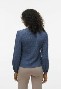 Top azzurro texture con maniche a sbuffo lunghe, caratterizzato da un collo rotondo e schemi delicati. Indossato con pantaloni beige.