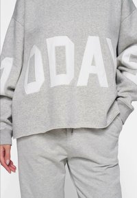 Jasnoszara bluza z oversize'owym krojem, z wyraźnym białym napisem "DZIŚ". Miękka tekstura, ściągacze przy mankietach i krótki krój u dołu.