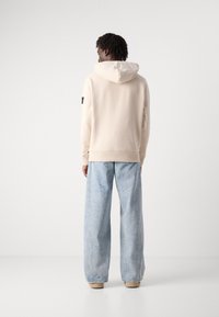 Felpa con cappuccio beige con una finitura testurizzata, abbinata a jeans a gamba larga blu chiaro. Presenta un'etichetta sul maniche sinistra.