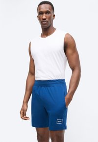 Άνδρας που φοράει λευκό αμάνικο πουκάμισο και μπλε σορτς Hugo, στέκεται με το ένα χέρι στην τσέπη, σε απλό φόντο.