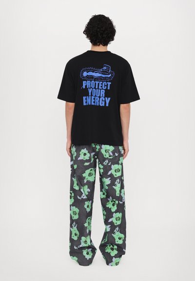 Schwarzes Baumwoll-T-Shirt mit blauer Schrift "PROTECT YOUR ENERGY" auf dem Rücken; kombiniert mit locker sitzenden grauen Hosen mit grünen Blumenmustern.