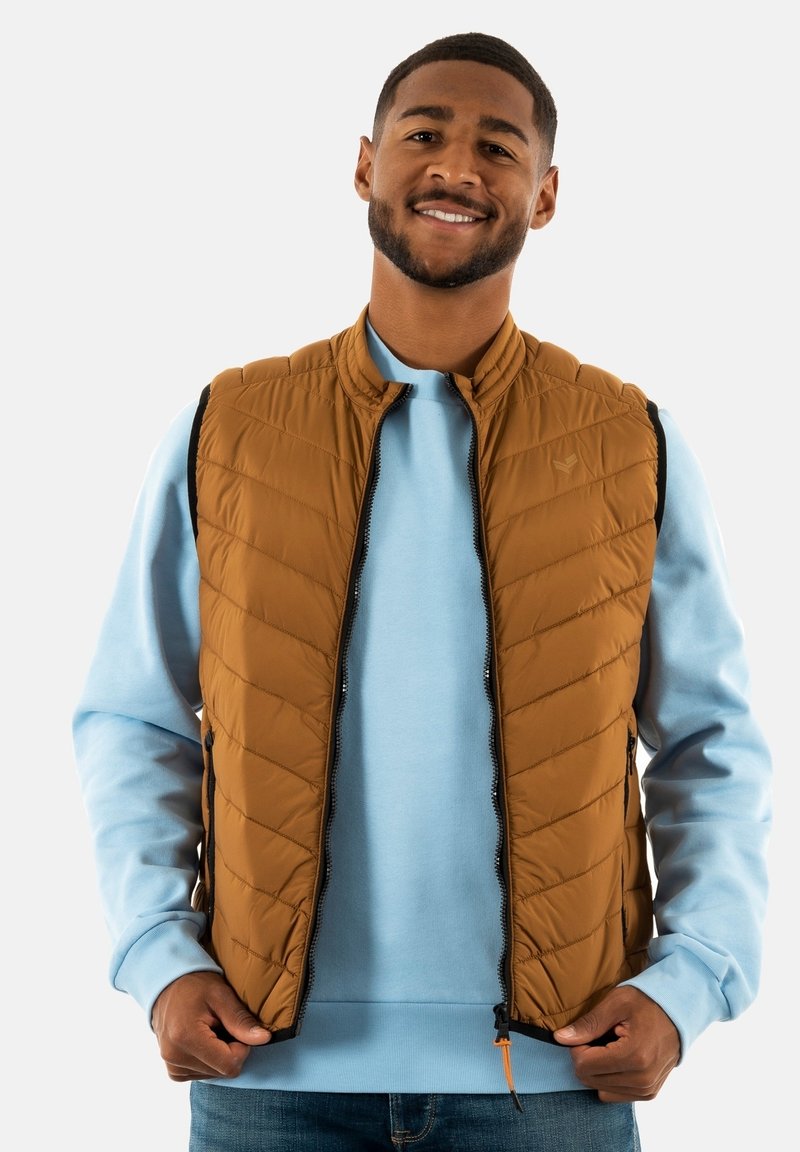Gilet trapuntato marrone con colletto alto, chiusura a zip e tasche laterali, indossato sopra una felpa azzurro chiaro. Tessuto testurizzato.