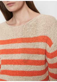 Kobieta w beżowym swetrze z włóczki z szerokimi, poziomymi pomarańczowymi paskami i okrągłym dekoltem, odsłaniającym szyję i górną część klatki piersiowej.