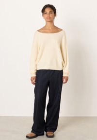 Kvinna stående i en gräddfärgad off-shoulder tröja, marinblå byxor med vita smala ränder och vida ben samt tvåfärgade skor i brunt och svart.