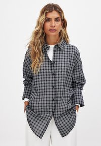Long-sleeve checkered shirt in black and gray with a button-down front, curved hem, and relaxed fit, layered over a white top.