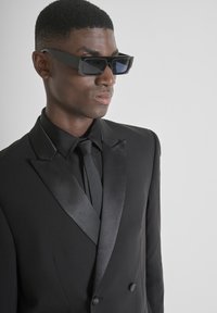 Esmoquin negro con solapas de satén y corbata a juego. Tela mate, ajuste estructurado, combinado con gafas de sol negras angulares con lentes oscuras.