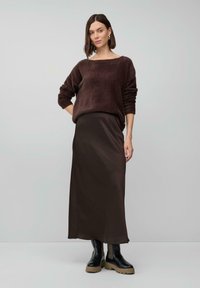 Femme portant un pull brun pelucheux, une longue jupe brune et des bottes noires avec des semelles beiges, se tenant devant un fond gris clair uniforme.