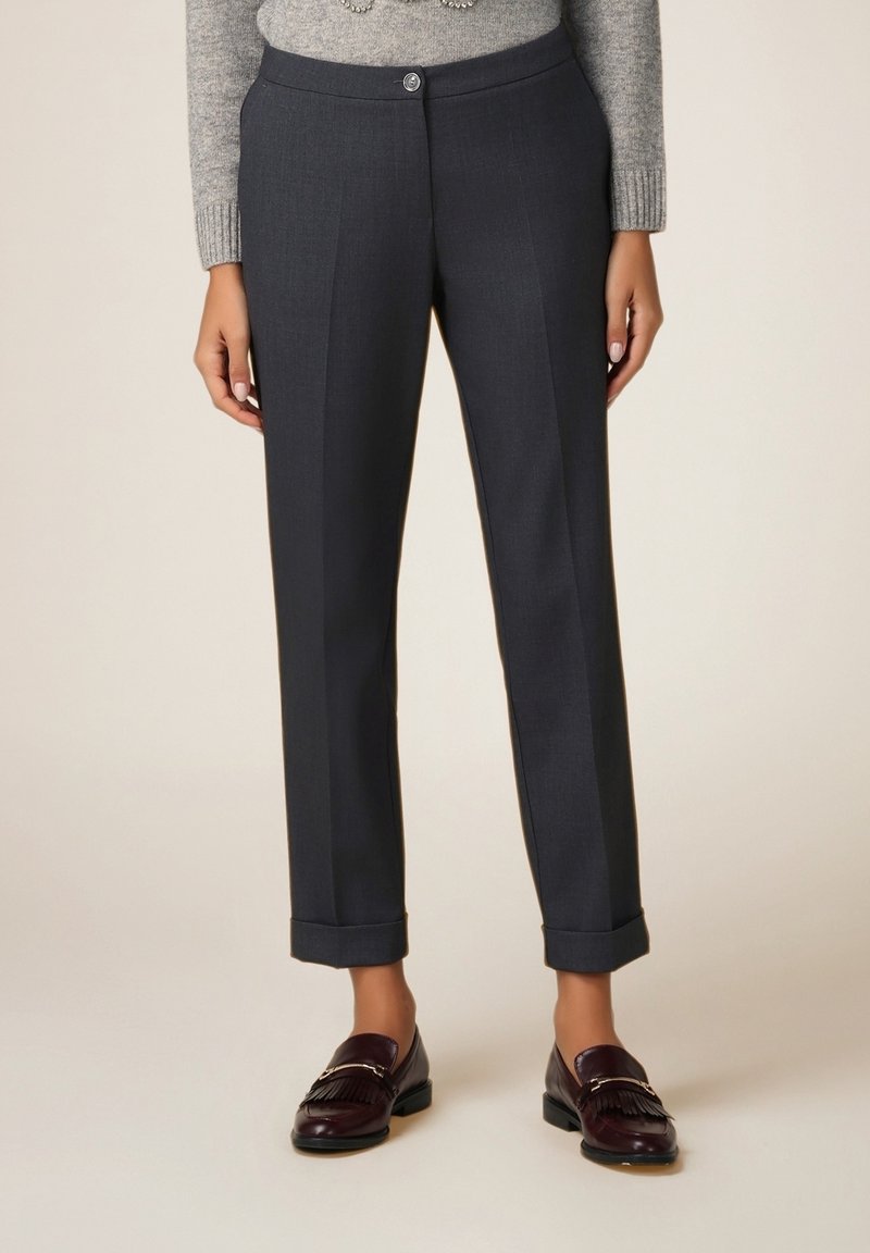 Femme portant un pantalon gris foncé sur mesure avec revers, un pull en maille gris et des mocassins en cuir marron foncé avec détails en franges.