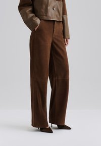 Pantaloni larghi in suede marrone con cuciture frontali e tasche laterali, abbinati a tacchi a punta marroni. Texture morbida, vestibilità su misura.