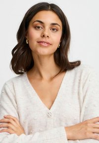 Ljus krämfärgad cardigan med texturpunkter, v-ringning och en enda knappknäppning. Modellen har mörkt hår och armarna i kors.
