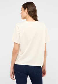 Kremowa koszulka z krótkim rękawem o luźnym kroju, okrągłym dekolcie i podwiniętych mankietach, w połączeniu z ciemnoniebieskimi dżinsami. Gładka faktura materiału.