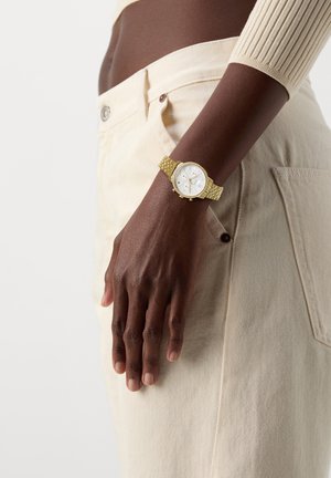 Mørkhudet person iført cremefarvede bukser og en lys sweater, der viser et guldur med hvid urskive på venstre håndled.