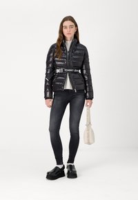 Svart puffervest med glidelås foran og logo-belte, kombinert med svarte skinny jeans og plattformklokker. Modell som holder en liten veske.