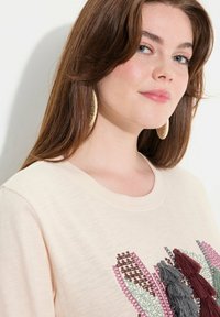 Helle beigefarbene T-Shirt mit Rundhalsausschnitt, das mit kunstvollen Stickereien in Burgunderrot, Grün und Grau verziert ist, akzentuiert durch dekorative Quasten.
