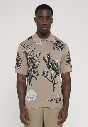 Mann trägt beige kurzärmeliges Poloshirt mit Blumenmuster und passende texturierte Hose, steht vor einfarbigem hellgrauem Hintergrund.