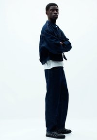 Jeansjakke og brede bukser i mørkeblå, parret med en hvid skjorte og sorte sneakers. Glatte teksturer, afslappet pasform.