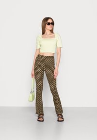 Top cropped verde claro com mangas bufantes, combinado com calças castanhas de boca de sino com um padrão geométrico. Acessórios incluem uma mala verde pálido e óculos de sol.