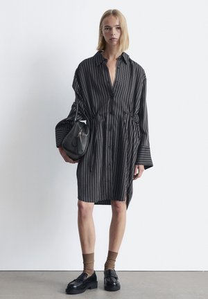 Black and white striped shirt dress with a loose fit, button-down front, and cinched waist, paired with a black handbag and loafers.