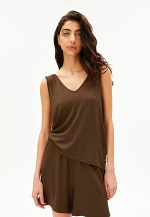 Vrouw met lang krullend haar, gekleed in een bruine mouwloze V-hals top en bijpassende losse short tegen een effen witte achtergrond.