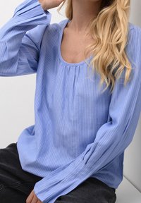 Blouse bleu clair à manches longues en tissu texturé, avec un col rond et de légères rayures verticales blanches. Coupe décontractée.